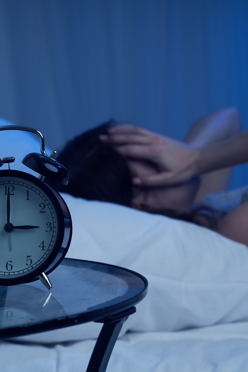 Woman lying in bed with head on pillow holding her face and an alarm clock in the foreground