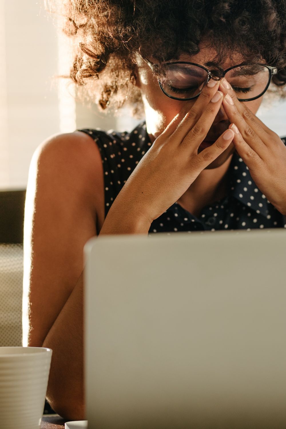 woman wearing glasses sitting at her desk in front of her computer with her hands in her face, with a cup of tea on the left