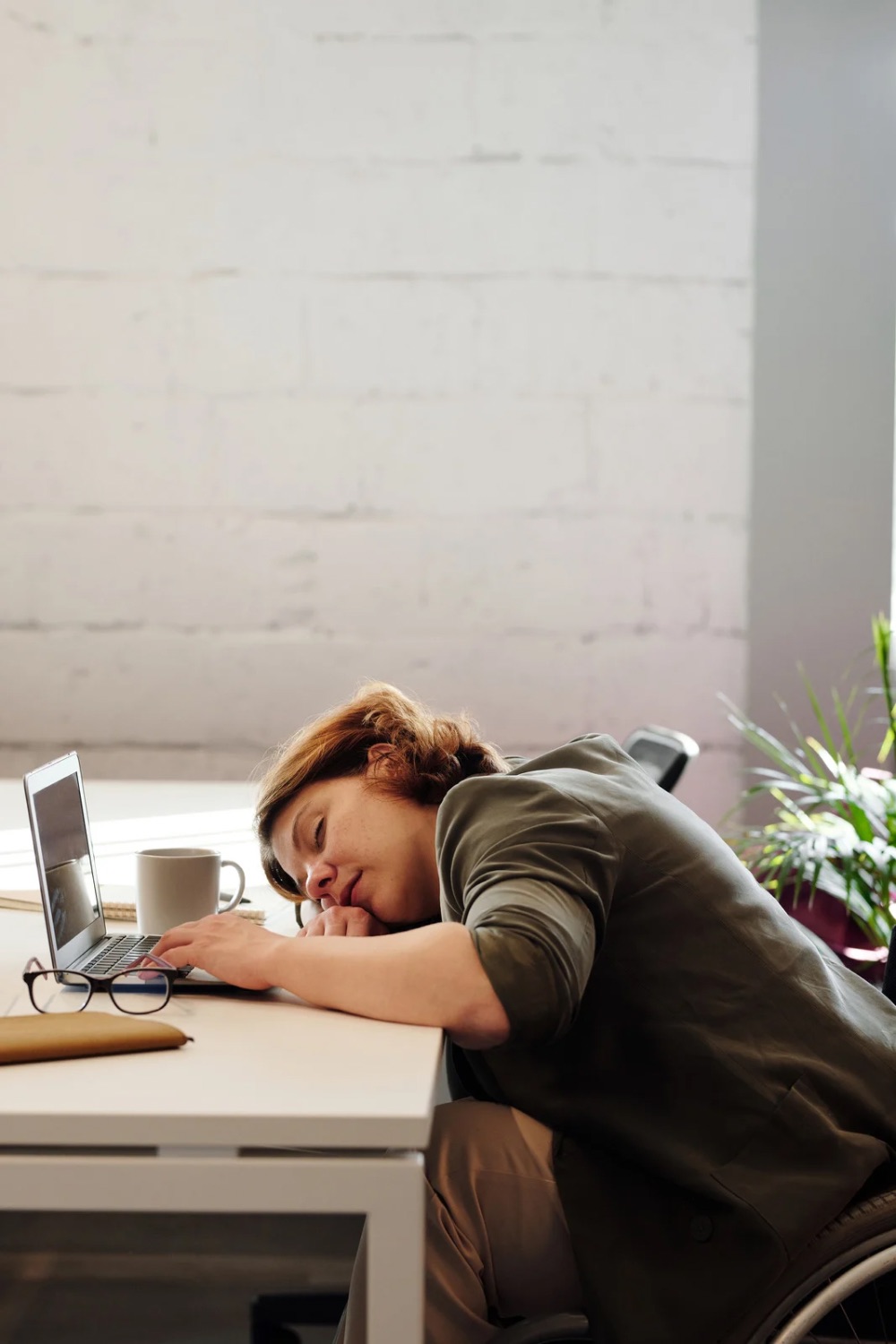 tired woman who has fallen asleep on her desk with a cup in her hand and her glasses and laptop on the table, with a house plant in the background