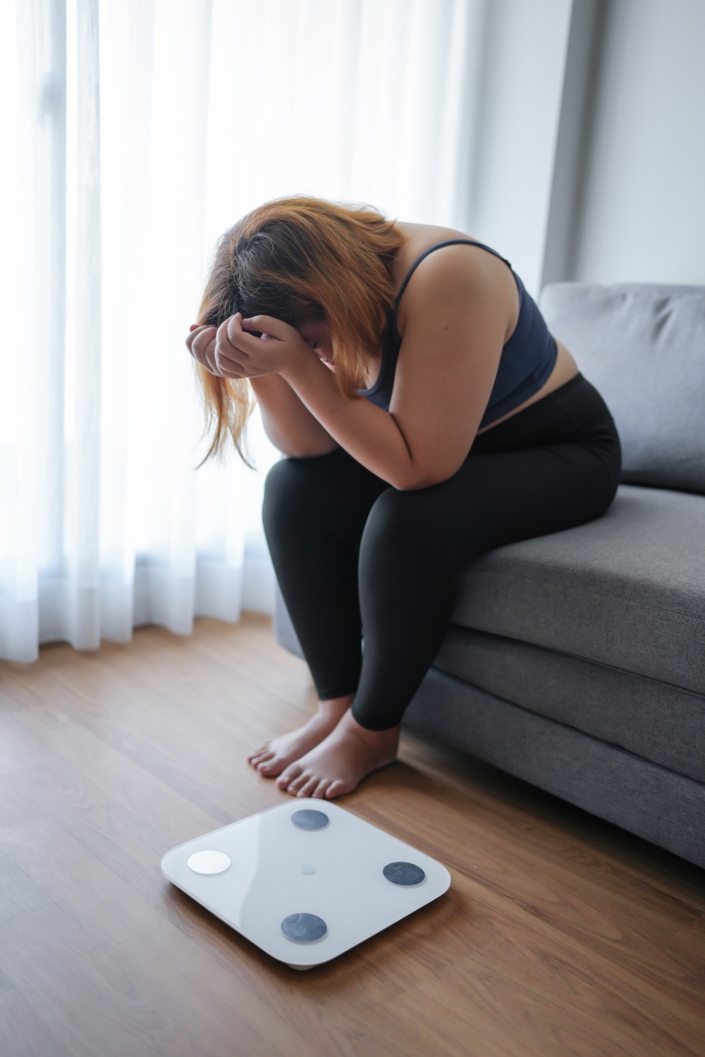 woman sitting on a sofa with her head in her hands looking down and her feet on a wooden floor, with bathroom scales in front of her, and a window with sunlight streaming in in the background