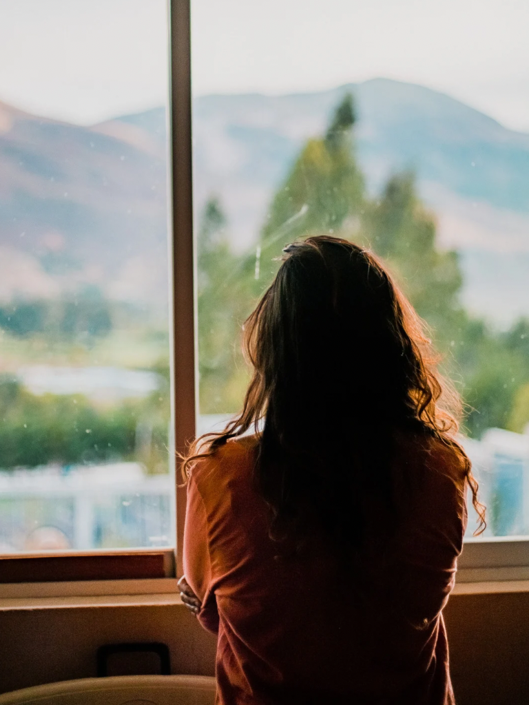 woman looking out of a window with mountains in the background, deep in thought about how she can resolver her low energy