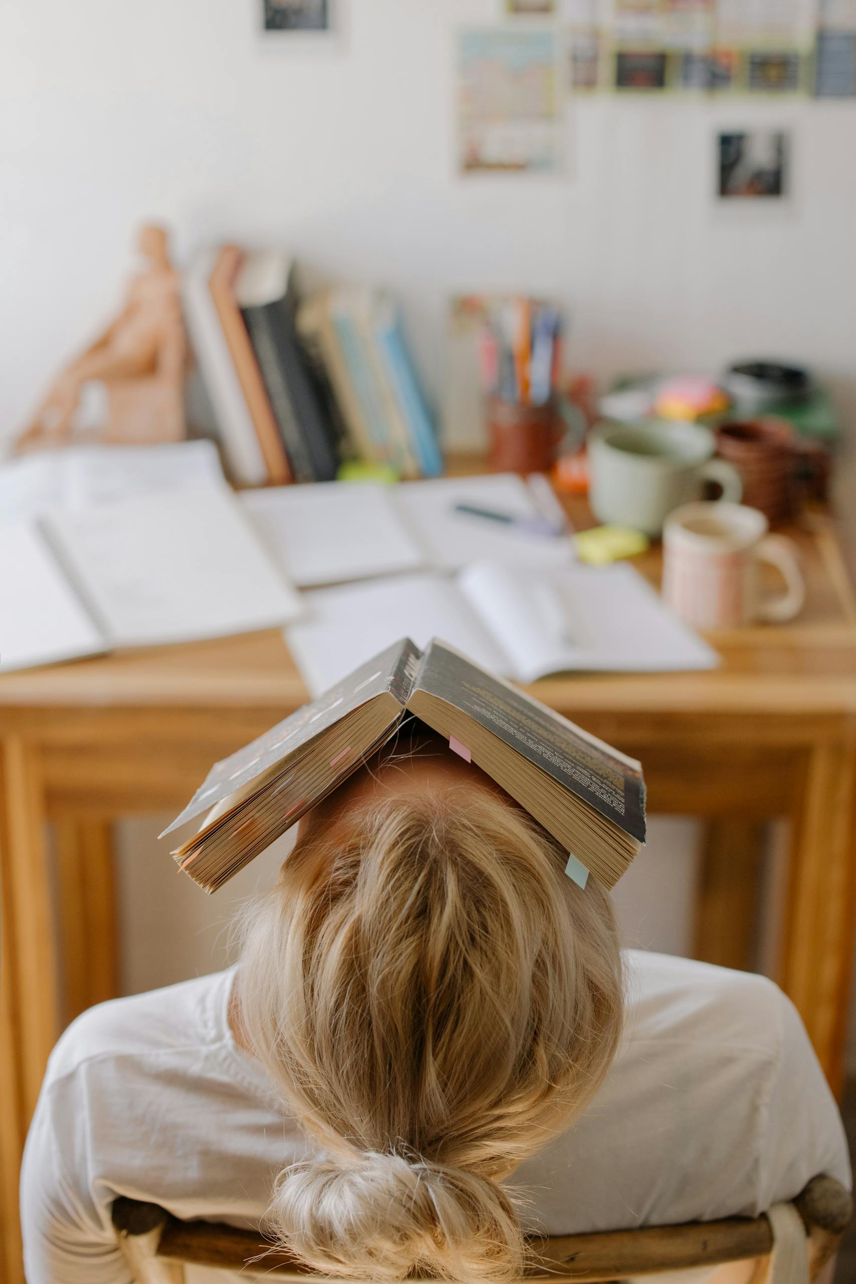 Woman feeling stress and exhaustion while working at a cluttered desk with an open book on their head.