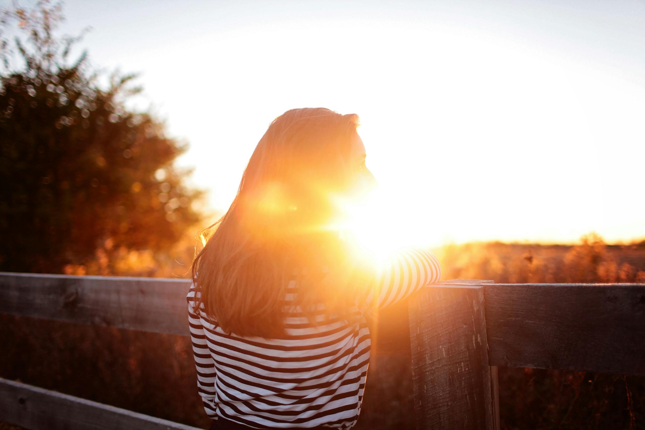A woman leans against a wooden fence, basking in the warm glow of a sunrise on a rural farm.